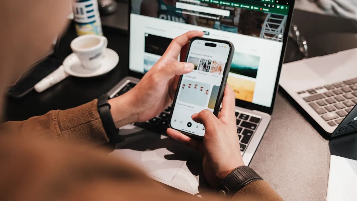 Eine Person am Schreibtisch hält ein Smartphone, davor ein offener Laptop.