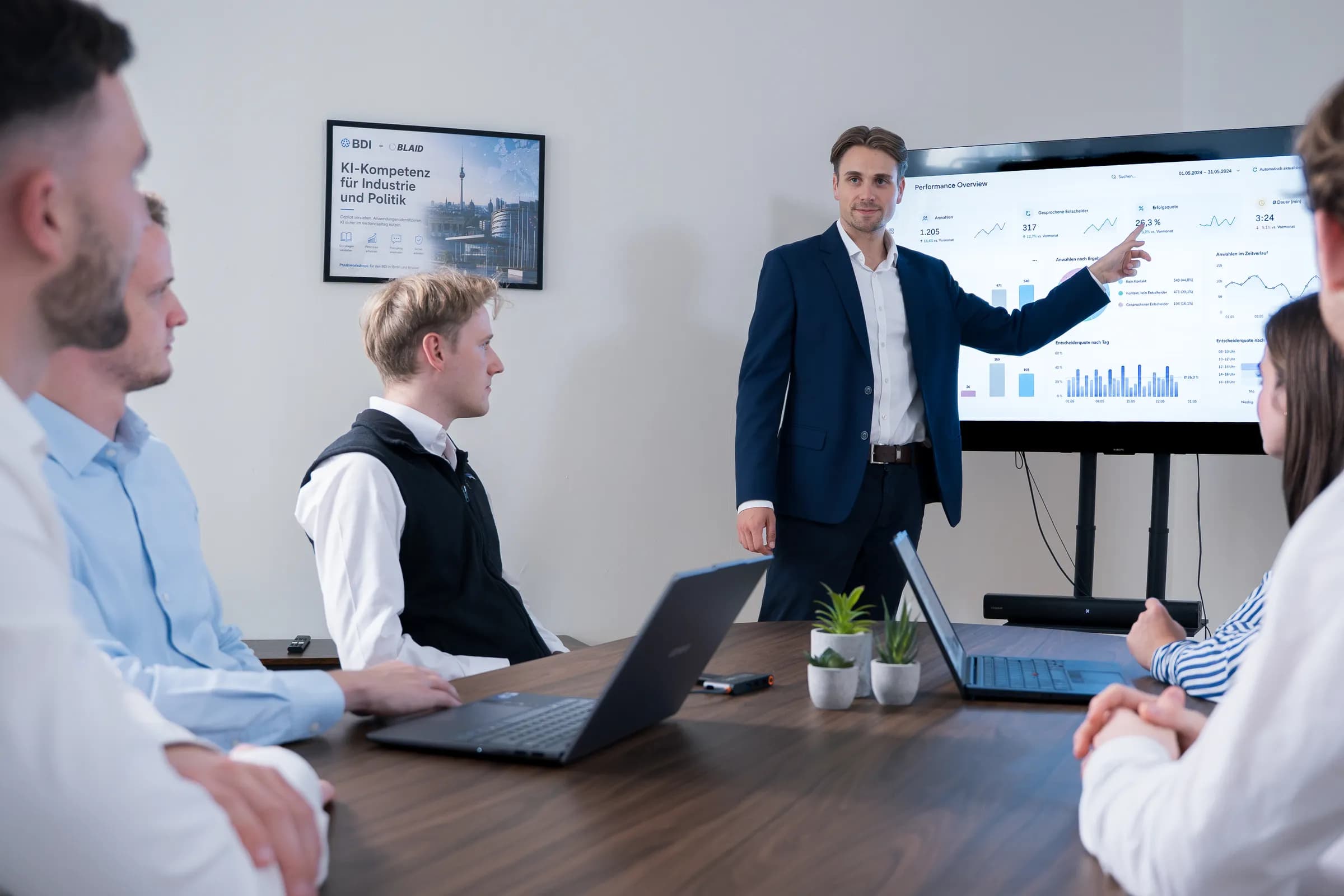 Ein BLAID-Workshop: Teilnehmende am Konferenztisch, davor ein Vortrag am Bildschirm mit einer Auswertung, an der Wand das Plakat „KI-Kompetenz für Industrie und Politik“ von BDI und BLAID.